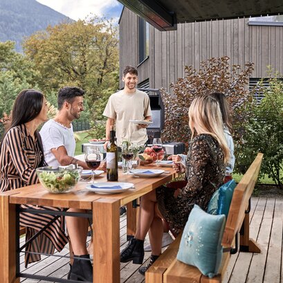 An afternoon barbecue is taking place in the garden and 4 people are already sitting on the Lago designer set and a man approaches them with a plate.