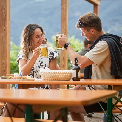 A couple clink two beer mugs together while sitting on classic beer tent sets on the terrace at the Guggerhof.